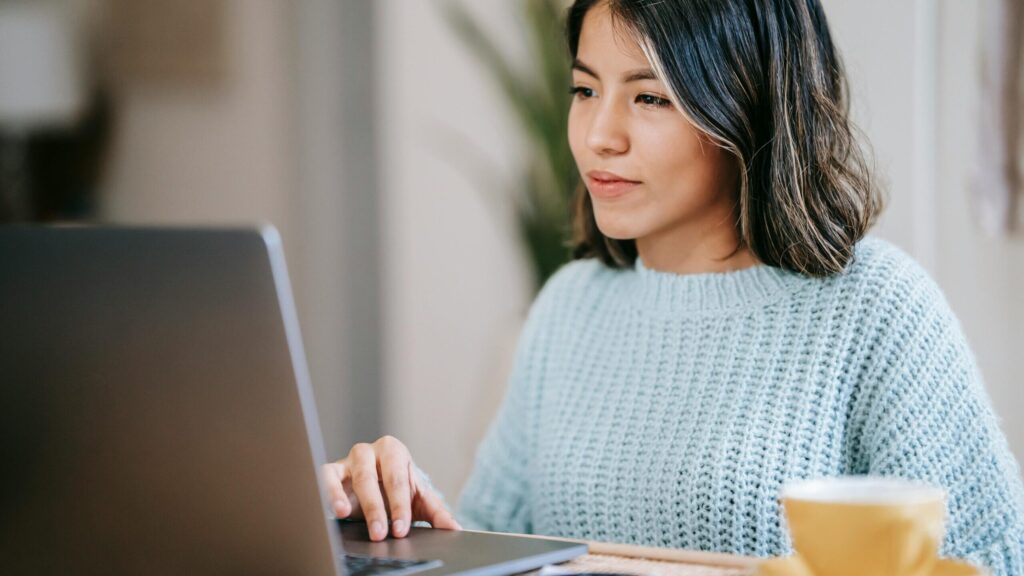 Confident young Latin American woman in casual clothes working remotely on netbook at table near mug with coffee in light apartment with plant with green leaves near wall